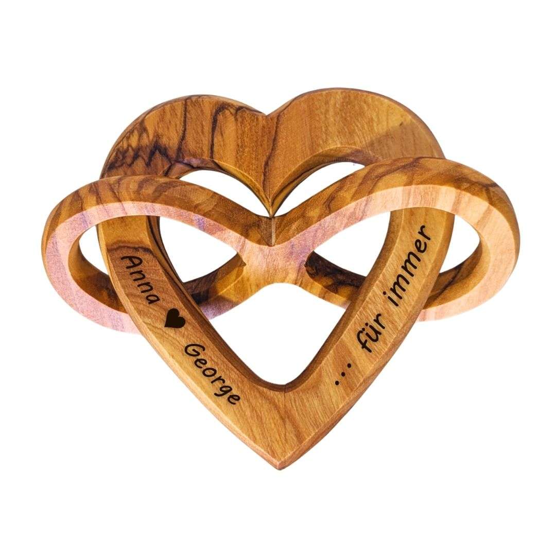 Interlocking wooden heart sculpture with two rings and engraved text reading 'Anna ♥ George ... für immer' indicating eternal love.