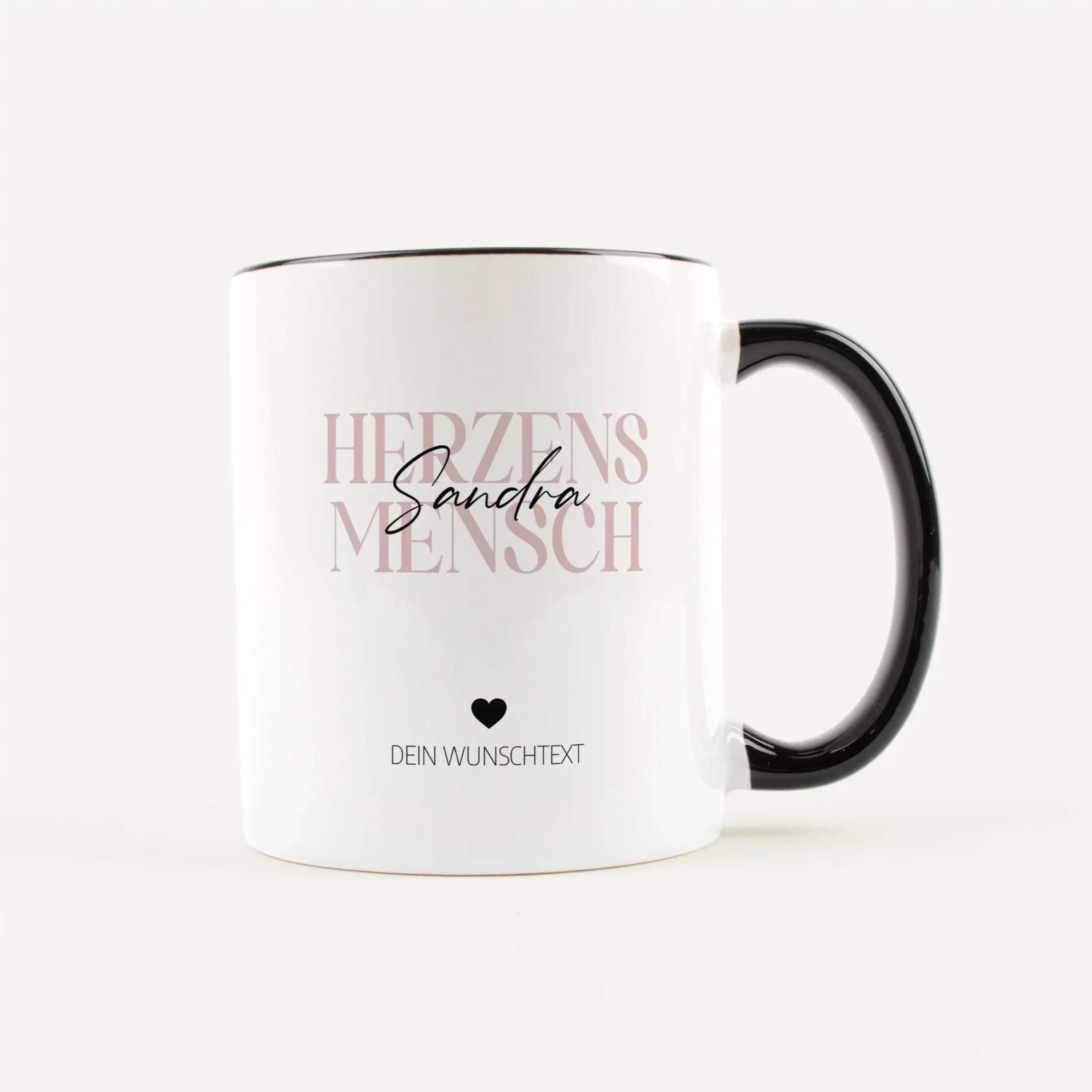 Close-up of a white mug with vertical pale-pink text reading 'HERZEN' and a black cursive 'Sandra' across; small heart icon and 'DEIN WUNSCHTEXT' at the bottom.