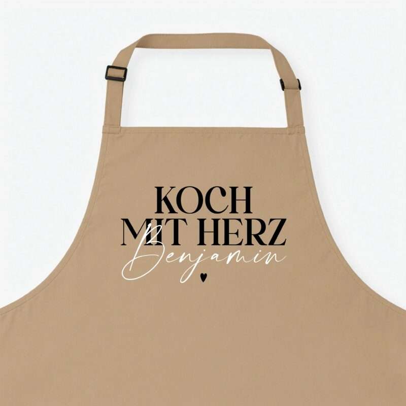 Beige apron with adjustable strap and bold black lettering across the front reading 'Koch mit Herz' (partial view).