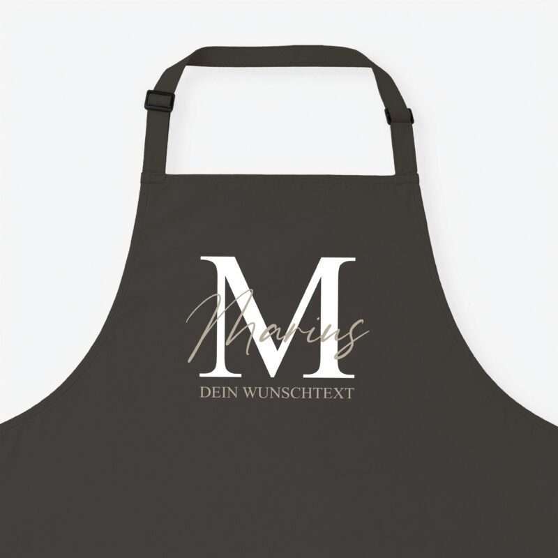 Dark apron with a large white 'MM' monogram and beige cursive text across the front, set on a light background (showing customizable text area).
