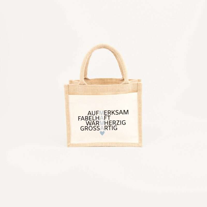 Beige burlap tote bag with handles, featuring stacked black German words and a small blue heart.